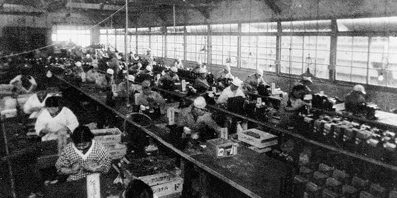 A historical photo of a Panasonic factory with rows of workers seated at long workbenches, engaged in detailed tasks.